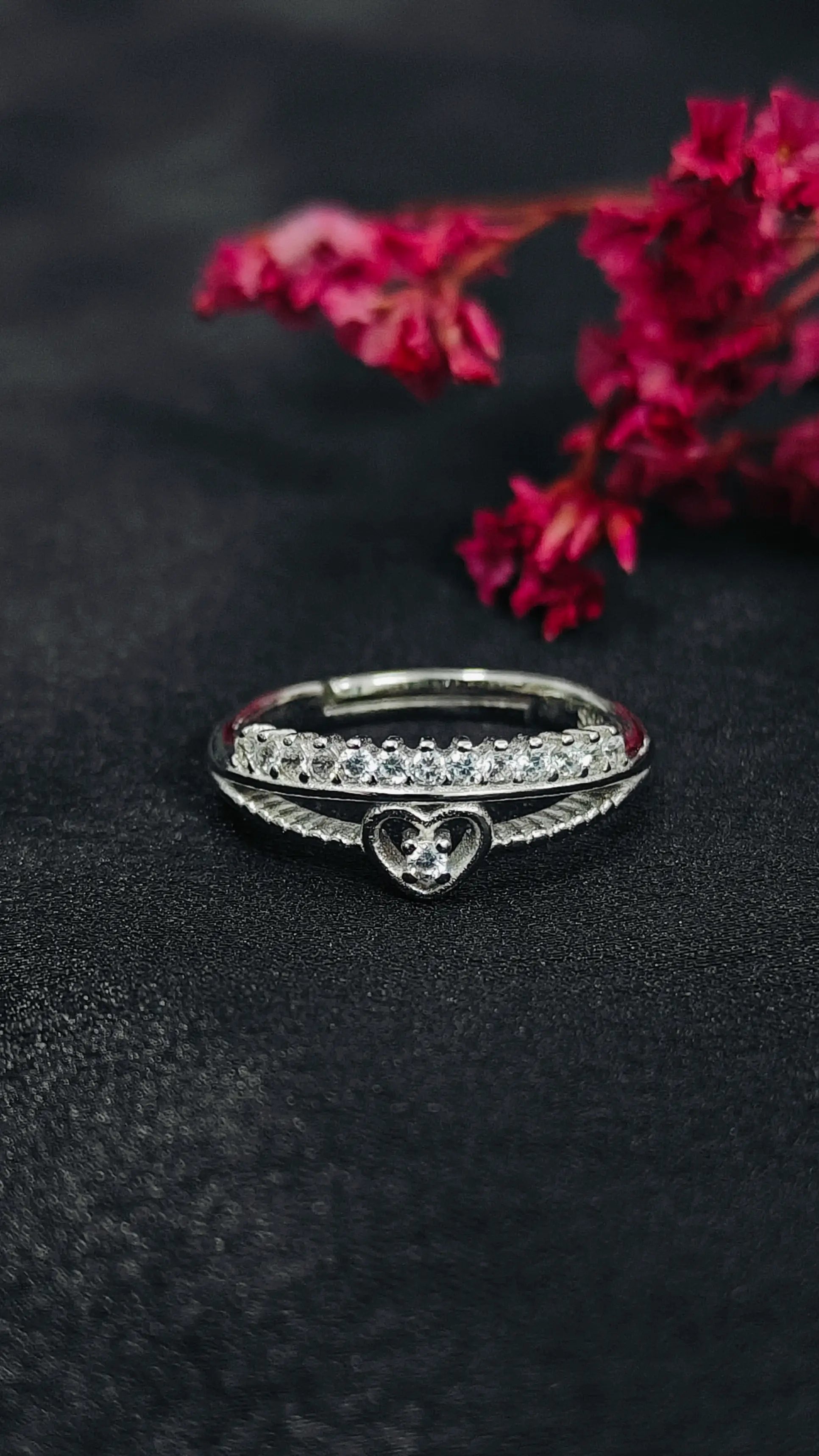 Silver ring with heart design on a black surface with pink flowers in the background